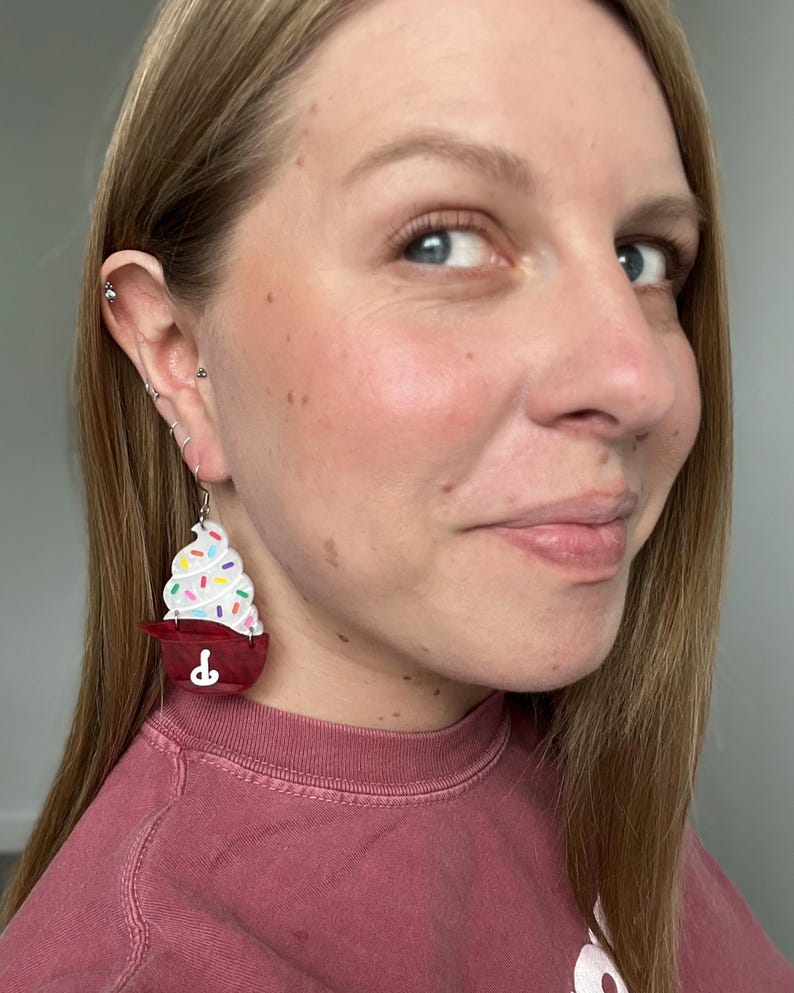 May include: A pair of novelty earrings shaped like cupcakes. The earrings feature a red cupcake holder with a white frosting swirl and colorful sprinkles. The earrings are hanging from silver hooks.
