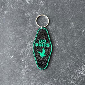 Op de afbeelding: Zwarte en groene motel sleutelhanger met een groen vogelsilhouet en de tekst "GO BIRDS" in groen.