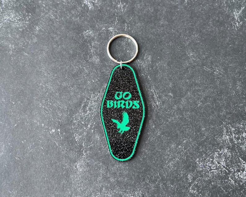 Op de afbeelding: Zwarte en groene motel sleutelhanger met een groen vogelsilhouet en de tekst "GO BIRDS" in groen.