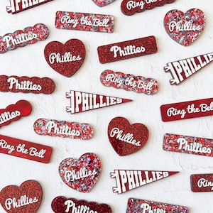 May include: A collection of red and white glitter-covered acrylic ornaments with the words "Phillies" and "Ring the Bell" on them.