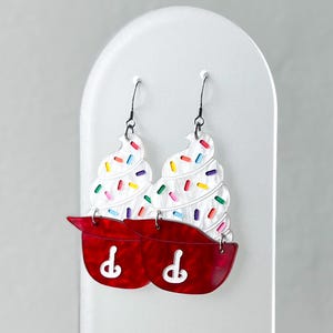 May include: A pair of whimsical earrings shaped like cupcakes. The earrings feature white frosting with colorful sprinkles atop a red cupcake liner. The earrings hang from black hooks and are displayed on a white stand.