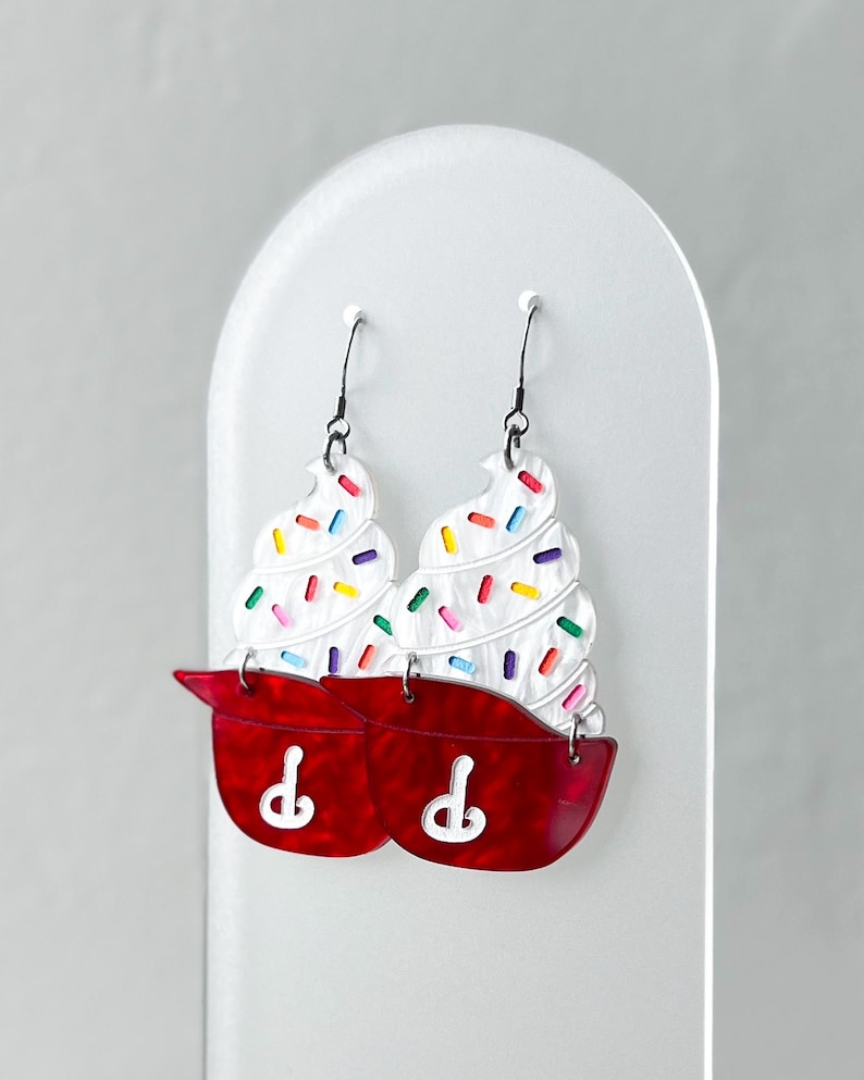 May include: A pair of whimsical earrings shaped like cupcakes. The earrings feature white frosting with colorful sprinkles atop a red cupcake liner. The earrings hang from black hooks and are displayed on a white stand.