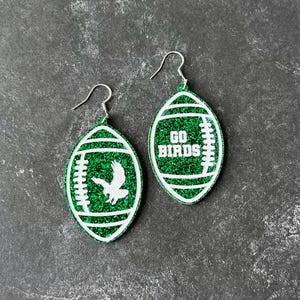 Może przedstawiać: Para zielonych błyszczących kolczyk&oacute;w w kształcie piłki nożnej z białymi błyszczącymi akcentami. Jeden kolczyk ma napis "GO BIRDS", a drugi ma białą sylwetkę ptaka.