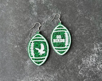 May include: A pair of green glitter football-shaped earrings with white glitter accents. One earring has the text "GO BIRDS" and the other has a white silhouette of a bird.