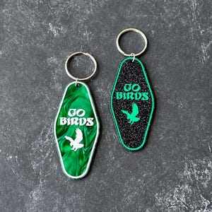 Op de afbeelding: Twee sleutelhangers met een motel-sleutelontwerp. De ene sleutelhanger is zwart met glitter en de andere is groen met een marmerpatroon. Beide sleutelhangers tonen de tekst "GO BIRDS" en een silhouet van een vogel.