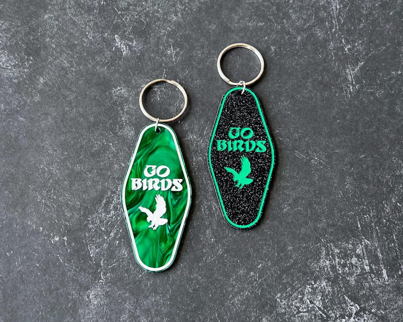 Op de afbeelding: Twee sleutelhangers met een motel-sleutelontwerp. De ene sleutelhanger is zwart met glitter en de andere is groen met een marmerpatroon. Beide sleutelhangers tonen de tekst "GO BIRDS" en een silhouet van een vogel.