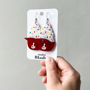 May include: A pair of novelty earrings shaped like red mixing bowls with white ice cream and colorful sprinkles. The earrings are on a white card with the text "studio Blade".