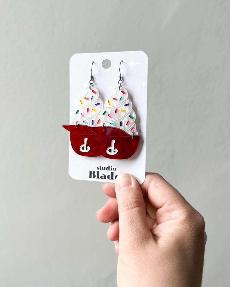 May include: A pair of novelty earrings shaped like red mixing bowls with white ice cream and colorful sprinkles. The earrings are on a white card with the text "studio Blade".