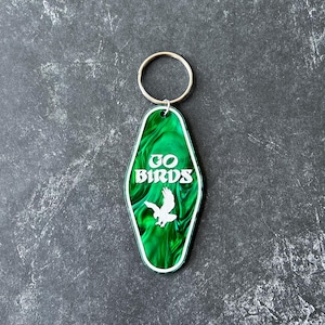 Op de afbeelding: Groene motel sleutelhanger met een wit silhouet van een vogel en de tekst "GO BIRDS".