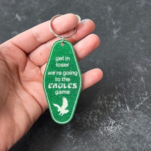 Op de afbeelding: Groene motel sleutelhanger met witte glitter en de tekst "Get in loser we're going to the Eagles game".