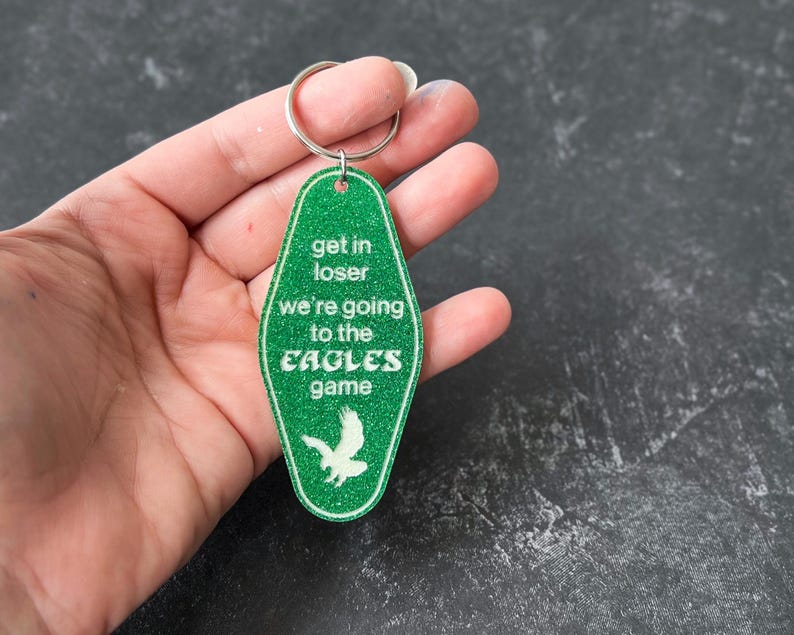 Op de afbeelding: Groene motel sleutelhanger met witte glitter en de tekst "Get in loser we're going to the Eagles game".