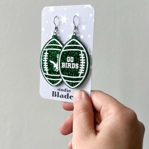 Może przedstawiać: Zielone błyszczące kolczyki w kształcie piłki nożnej z białym napisem "GO BIRDS". Kolczyki znajdują się na białej kartce z napisem "studio Blade".