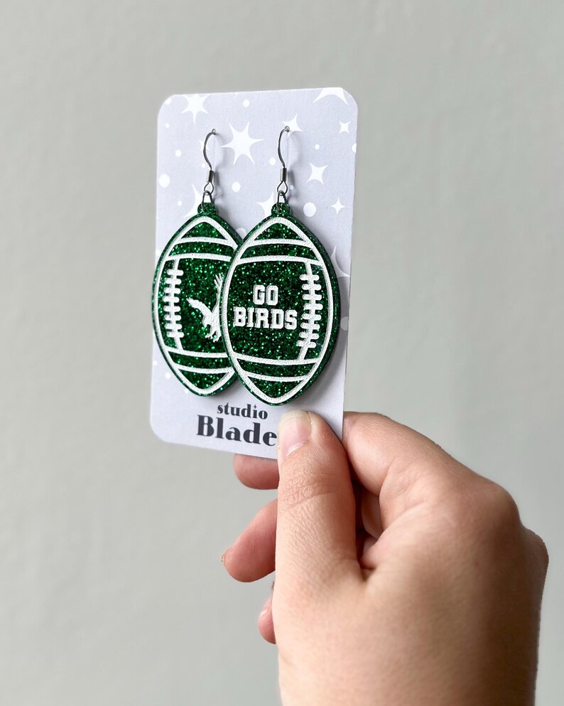 Może przedstawiać: Zielone błyszczące kolczyki w kształcie piłki nożnej z białym napisem "GO BIRDS". Kolczyki znajdują się na białej kartce z napisem "studio Blade".