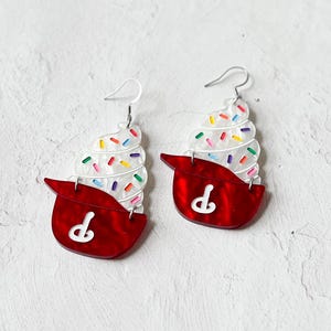 May include: A pair of novelty earrings shaped like ice cream cones. The earrings feature a red base with a white design and a white ice cream swirl topped with colorful sprinkles. The earrings have silver-colored hooks.