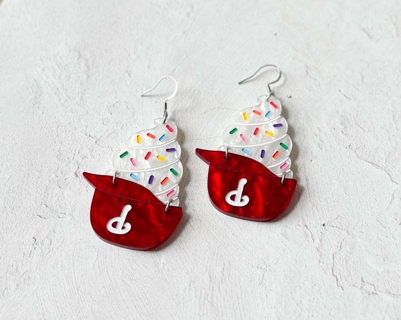May include: A pair of novelty earrings shaped like ice cream cones. The earrings feature a red base with a white design and a white ice cream swirl topped with colorful sprinkles. The earrings have silver-colored hooks.