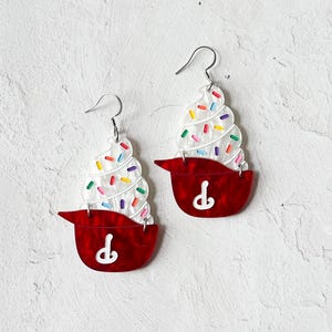 May include: A pair of novelty earrings shaped like cupcakes. Each earring features a red base with a white swirl of frosting and colorful sprinkles. The earrings have silver-colored fish hook ear wires.