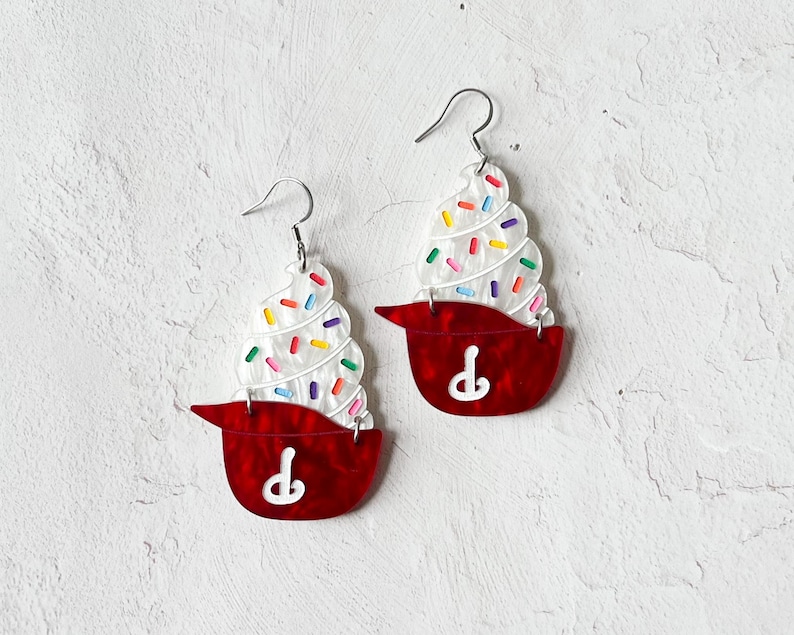May include: A pair of novelty earrings shaped like cupcakes. Each earring features a red base with a white swirl of frosting and colorful sprinkles. The earrings have silver-colored fish hook ear wires.