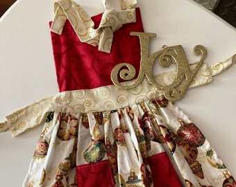 May include: A red and white Christmas-themed apron with a gold glitter "Joy" sign. The apron features a white background with a pattern of red and gold ornaments. The apron has a red pocket and a red top with a gold swirl pattern.