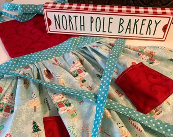 May include: A turquoise and white apron with a red pocket and a white polka dot trim. The apron has a gingerbread house pattern and the text "North Pole Bakery" is on a red and white sign.