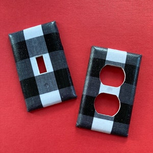May include: Two white outlet covers with black and white buffalo plaid pattern. One cover has a single light switch opening and the other has a standard outlet opening.