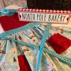 May include: A turquoise and white apron with a red pocket and a white polka dot trim. The apron has a gingerbread house pattern and the text "North Pole Bakery" is on a red and white sign.