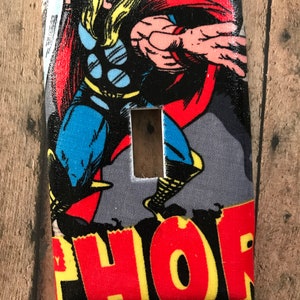 May include: A fabric light switch cover featuring a comic book illustration of Thor. The design includes Thor in a dynamic pose with a red cape, blue and yellow accents, and the word "THOR" in bold letters.