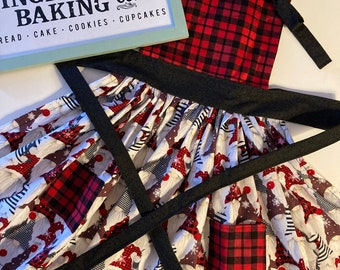 May include: A red and black plaid apron with a black adjustable strap and a pocket. The apron has a white background with a pattern of gnomes wearing red and white hats. The apron has the text "Gingerbread Baking Co." on it.