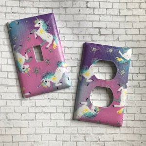 May include: Two light switch covers with a pink, purple, and blue ombre background. The covers feature a unicorn pattern with glitter accents.