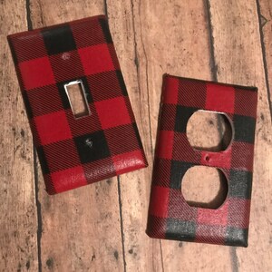 May include: Two red and black buffalo plaid light switch and outlet covers. The switch cover has a single switch opening and the outlet cover has two openings.