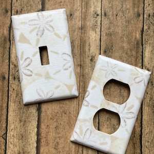 May include: Two white light switch and outlet covers with a beige floral pattern. The covers are on a wood background.