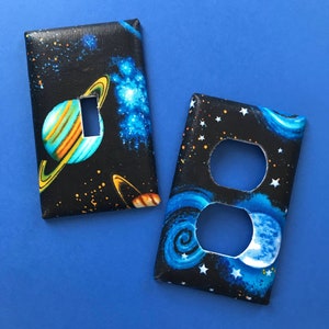 May include: Two decorative light switch covers with a space theme. One cover features a planet with rings and a galaxy background. The other cover features a swirling galaxy with stars and a crescent moon.