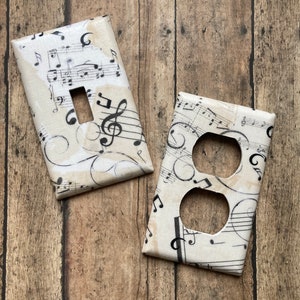 May include: Two decorative light switch covers with a musical theme. One cover has a single switch opening, and the other has two outlet openings. Both are covered in a cream-colored fabric printed with black musical notes and staff lines.
