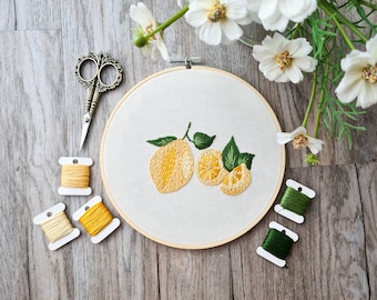 May include: A close-up of an embroidery hoop with a lemon design. The hoop is on a wooden surface with spools of yellow and green thread.