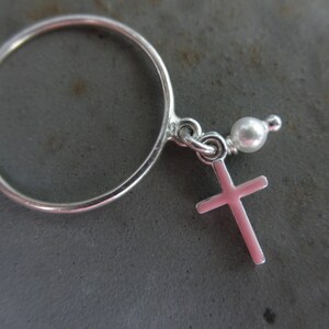 May include: A silver ring with a small pink cross charm and a white pearl bead. The ring is simple and elegant, with a delicate design. The cross is a classic religious symbol.