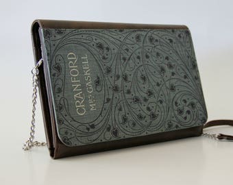 Könnte beinhalten: Eine rechteckige Clutch mit Buchcover-Design. Die Vorderseite zeigt ein graues Blumenmuster und den Titel "Cranford" von Mrs. Gaskell. Die Kanten sind braun, und sie hat eine silberne Kette und einen braunen Lederriemen.