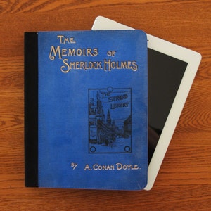Könnte beinhalten: Ein blauer Buchumschlag mit goldenen Lettern, auf dem "The Memoirs of Sherlock Holmes" und "By A. Conan Doyle" steht. Der Buchumschlag zeigt außerdem eine Schwarzweißillustration einer Straßenszene mit dem Text "The Strand Library".