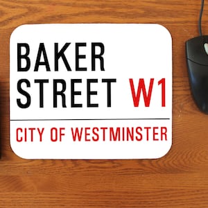 May include: A white rectangular mousepad with rounded corners, featuring the text "BAKER STREET W1 CITY OF WESTMINSTER" in black and red. A black keyboard and mouse are on a wooden surface.
