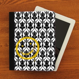 May include: Black and white tablet case with a black border. The case features a repeating pattern of stylized black flowers on a white background. A yellow smiley face is spray painted over the pattern.