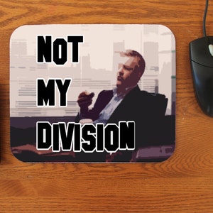 May include: Rectangular mousepad with the text "NOT MY DIVISION" in bold, black letters. The mousepad features an image of a man in a suit, seated and holding a cigarette. A black computer mouse and keyboard are also visible.