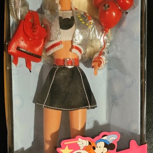 K&ouml;nnte beinhalten: Eine Barbie-Puppe tr&auml;gt eine silberne Jacke mit roten Verzierungen, einen schwarzen Rock und ein wei&szlig;es Hemd mit einer schwarzen Mickey-Maus-Silhouette. Sie h&auml;lt einen roten Mickey-Maus-Ballon und einen roten Rucksack mit der Nummer 25 darauf. Die Puppe befindet sich in einer rosa Schachtel mit dem Text "Walt Disney World Barbie" und "Special Edition" darauf.