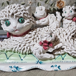 May include: A white ceramic cat ornament with three kittens. The mother cat has green eyes and a red collar. The kittens also have red bows. The ornament sits on a green and white base with blue accents.