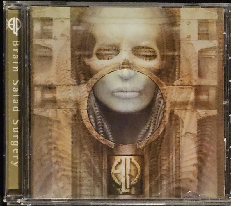 K&ouml;nnte beinhalten: CD-H&uuml;lle mit dem Albumcover von "Brain Salad Surgery". Das Artwork zeigt ein surrealistisches, sepiafarbenes Bild eines Gesichts mit kreisf&ouml;rmiger &Ouml;ffnung, mechanischen Elementen und dem Logo der Band.