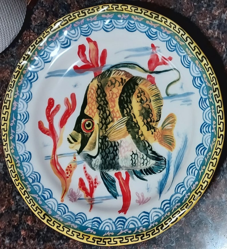 May include: A white ceramic plate with a blue and yellow border. The plate features a colorful illustration of a fish with red coral and seaweed.