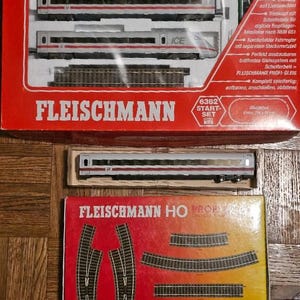 Puede incluir: Una caja roja y blanca con el texto "Fleischmann Start-Set Intercityexpress ICE 2" y un set de tren modelo en su interior. El tren es blanco con rayas rojas y el texto "ICE" en el lateral. La caja también incluye una caja más pequeña con el texto "Fleischmann HO Profi" y un conjunto de vías de tren.