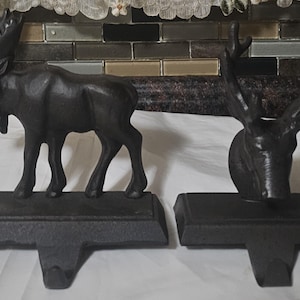 May include: Two dark brown cast iron stocking holders. One is a standing moose, the other is a deer head. Both have a rectangular base with a hook for hanging stockings. The moose is approximately 20 cm tall.