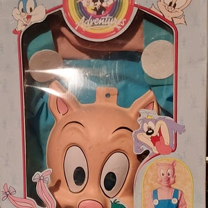 Puede incluir: Un conjunto de disfraz de Tiny Toon Adventures que incluye un mono rosa y azul, una máscara de cerdo de plástico y una ilustración del personaje de Hampton el cerdo. La caja incluye el texto "Tiny Toon Adventures" y "Hampton".