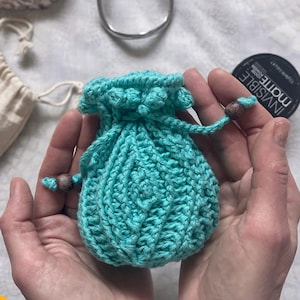 May include: A turquoise crochet drawstring pouch held in open hands. Other items are visible, including a silver bracelet, a small round blue and gold container, and a small beige bag. The words "written charted video" are in a yellow circle.