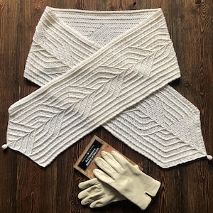 May include: Cream-colored, hand-crocheted scarf with a geometric pattern, lying on a wooden surface. The scarf has a V-shape and is paired with a pair of light-colored gloves and a book titled "All I Crossed A Bridge of Dreams".