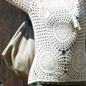May include: Cream-colored crochet top with long sleeves and circular patterns. The open-weave design reveals a glimpse of the undergarment. The top has a relaxed fit and a tassel detail at the neckline. The background is a dark wooden surface.