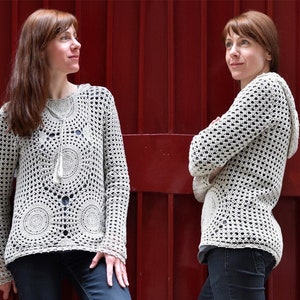 May include: A cream-colored, open-weave crochet sweater with a hood and long sleeves. The sweater features circular patterns on the front and a drawstring neckline. The model is wearing dark blue jeans. The background is a solid red.